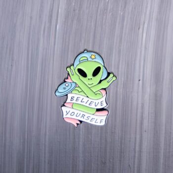 Alien believe yourself enamel pin green with blue hat. These enamel pins are made with metal. These enamel pins are colorful, with a black backing.