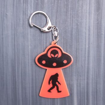 Bigfoot Abduction Neon Orange Bag Tag with black ship and black backing. Acrylic designed charm with a swivel lobster snap hook in high quality metal alloy.