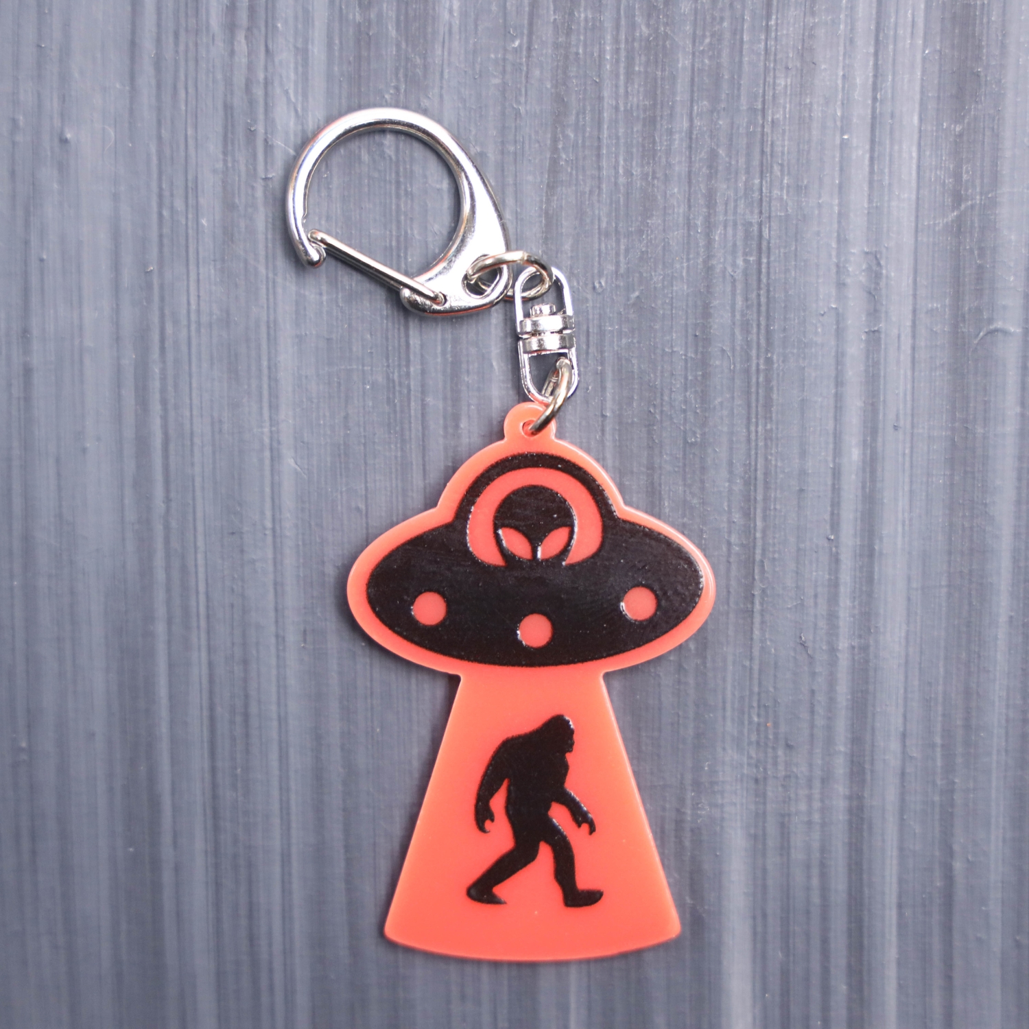 Bigfoot Abduction Neon Orange Bag Tag with black ship and black backing. Acrylic designed charm with a swivel lobster snap hook in high quality metal alloy.