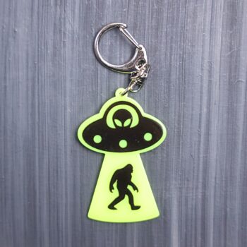 Bigfoot Abduction Neon Yellow Bag Tag with black ship and black backing. Acrylic designed charm with a swivel lobster snap hook in high quality metal alloy.
