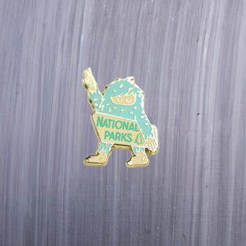 Bigfoot National Parks Enamel Pin in green and white with bigfoot holding up a peace sign. These enamel pins are made with metal. These enamel pins are colorful, with a gold colored backing.