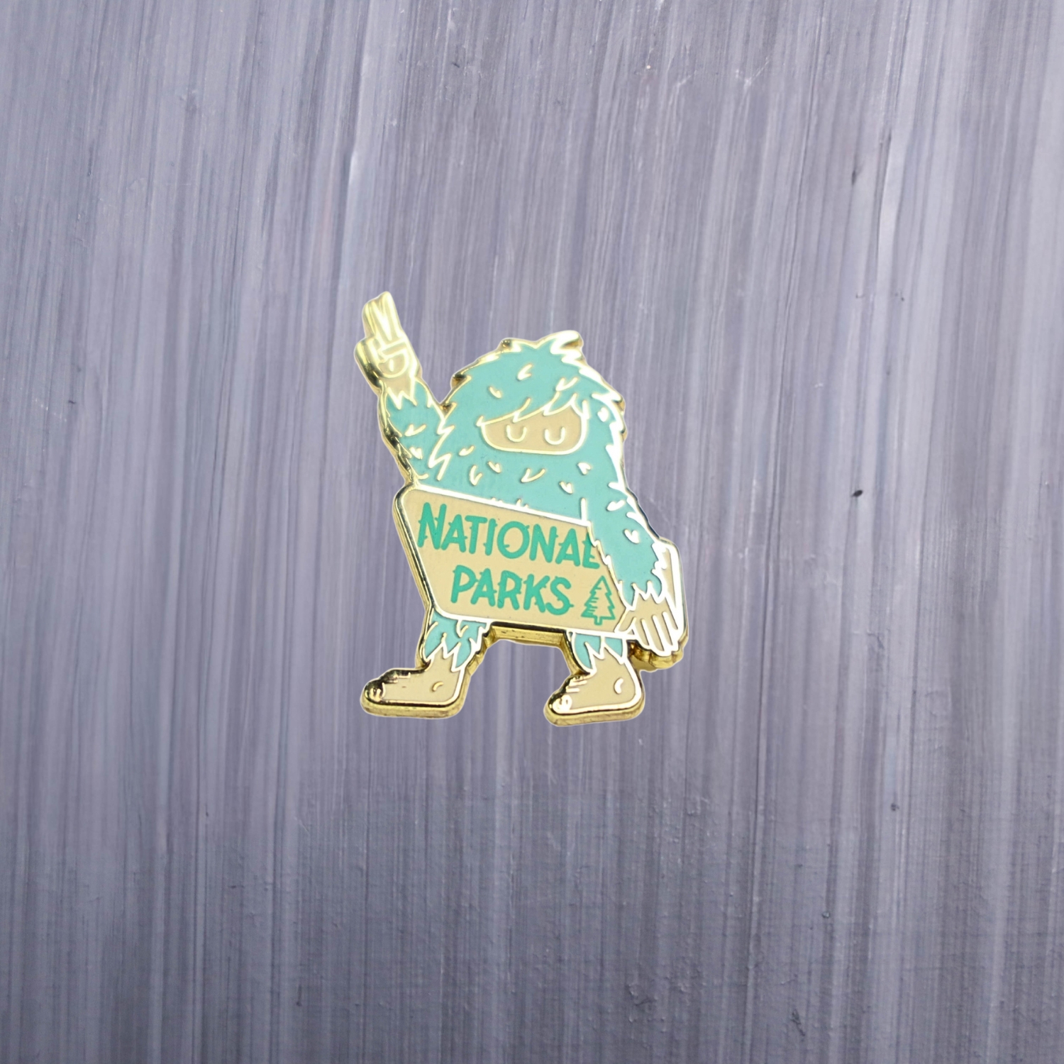 Bigfoot National Parks Enamel Pin in green and white with bigfoot holding up a peace sign. These enamel pins are made with metal. These enamel pins are colorful, with a gold colored backing.