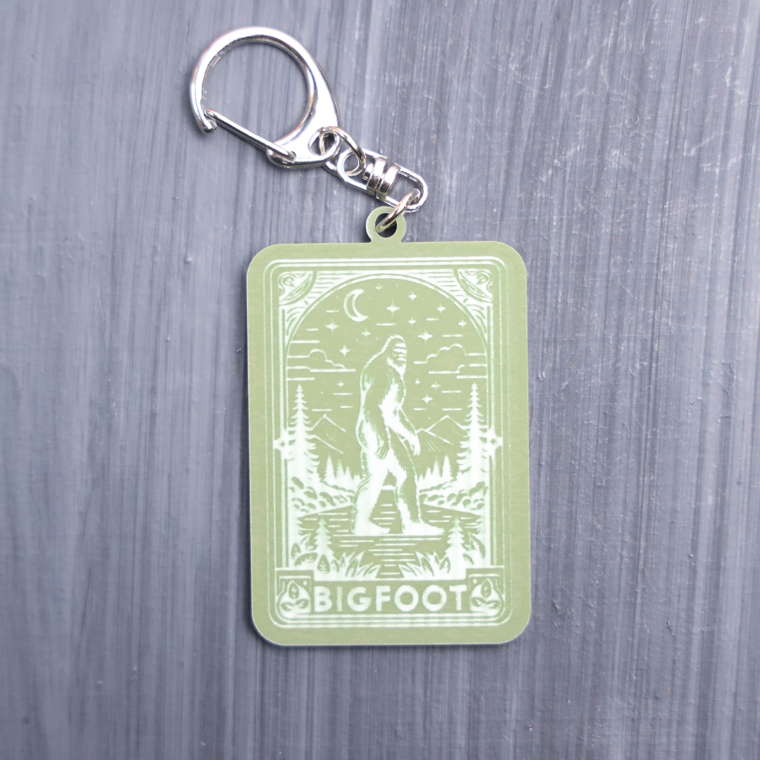 Bigfoot tarot card bag tag in green with trees, moon, and mountains. Acrylic designed charm with a swivel lobster snap hook in high quality metal alloy.