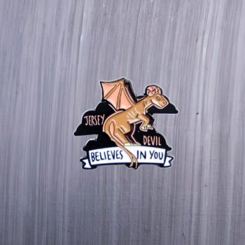 Jersey Devil Believes In You Enamel Pin in brown, pink, and black. These enamel pins are made with metal. These enamel pins are colorful, with a black backing.