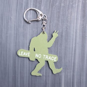 Leave No Trace Bigfoot Bag Tag in green with snow board. Acrylic designed charm with a swivel lobster snap hook in high quality metal alloy.