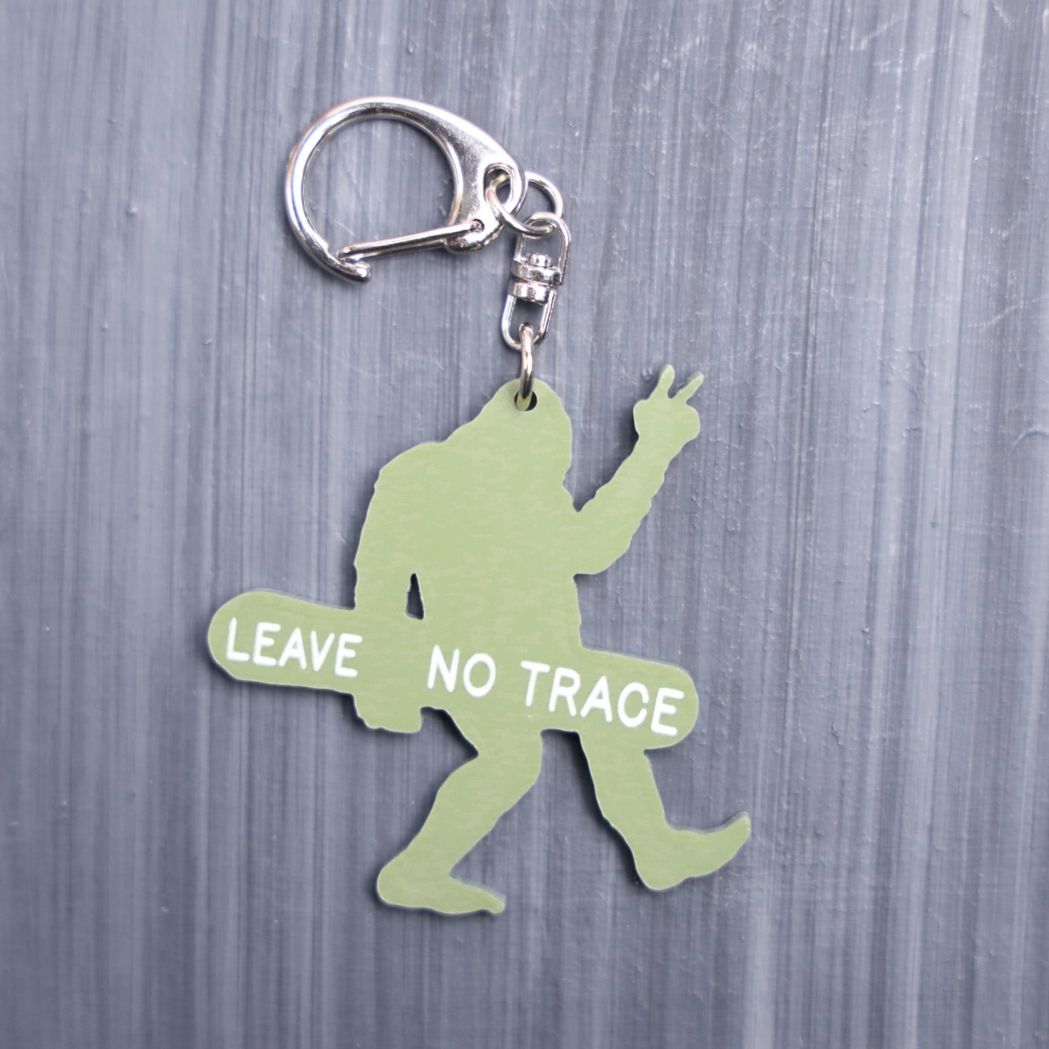 Leave No Trace Bigfoot Bag Tag in green with snow board. Acrylic designed charm with a swivel lobster snap hook in high quality metal alloy.