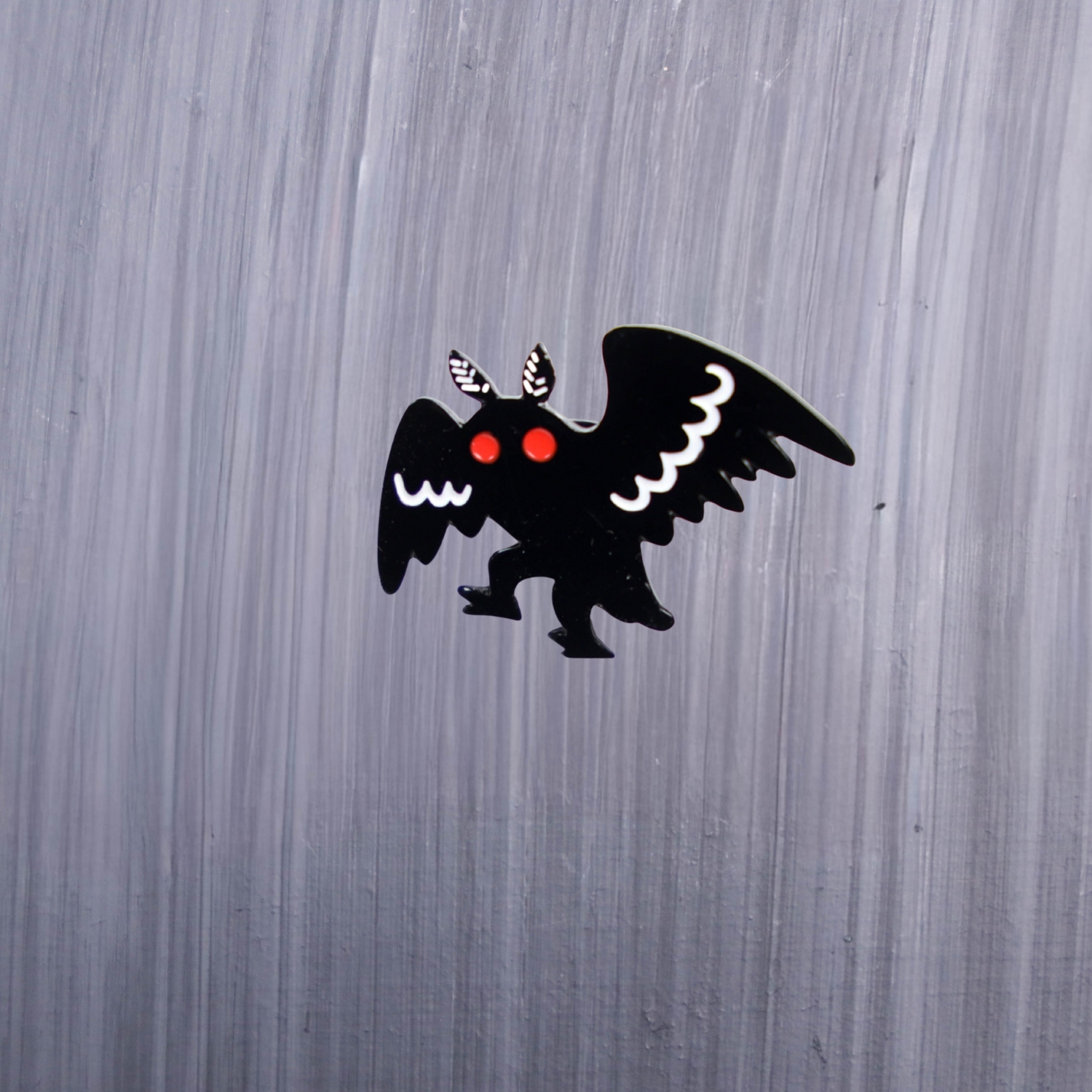 Mothman Walking Enamel Pin with black and white wings and red eyes. These enamel pins are made with metal. These enamel pins are colorful, with a black backing.