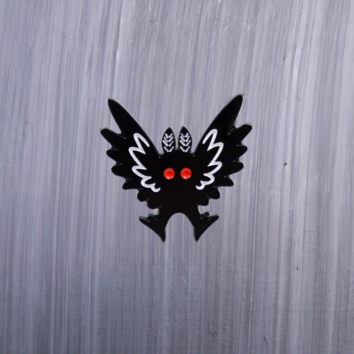 Mothman Wings Up Enamel Pin with black and white wings up and red eyes. These enamel pins are made with metal. These enamel pins are colorful, with a black backing.