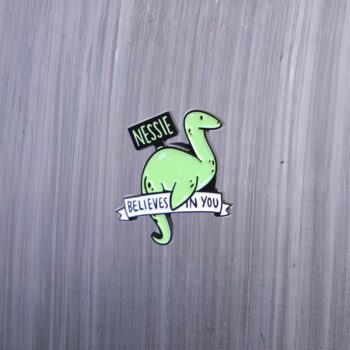 Nessie Believes In You Enamel Pin in green with sign. These enamel pins are made with metal. These enamel pins are colorful, with a black backing.