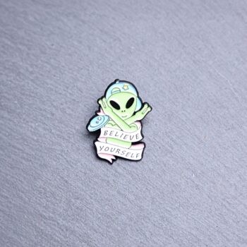 Alien believe yourself enamel pin green with blue hat. These enamel pins are made with metal. These enamel pins are colorful, with a black backing.