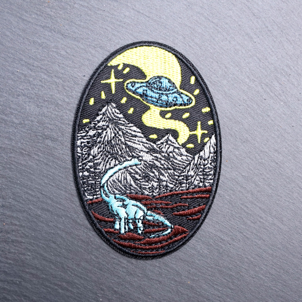 Alien Dinosaur Patch with spaceship, mountains, and dinosaur. Beautifully embroidered design with an adhesive for easy application to your favorite bag or article of clothing.