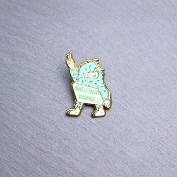Bigfoot National Parks Enamel Pin in green and white with bigfoot holding up a peace sign. These enamel pins are made with metal. These enamel pins are colorful, with a gold colored backing.