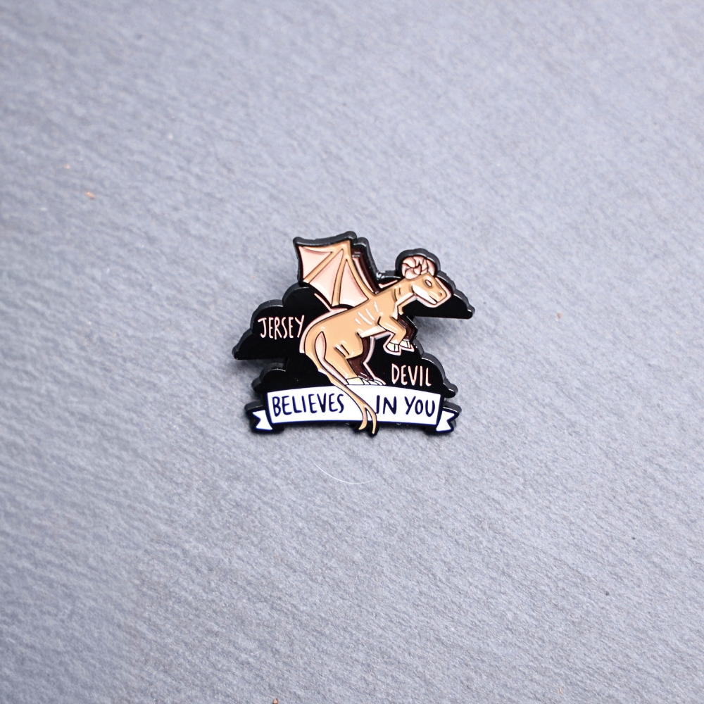 Jersey Devil Believes In You Enamel Pin in brown, pink, and black. These enamel pins are made with metal. These enamel pins are colorful, with a black backing.
