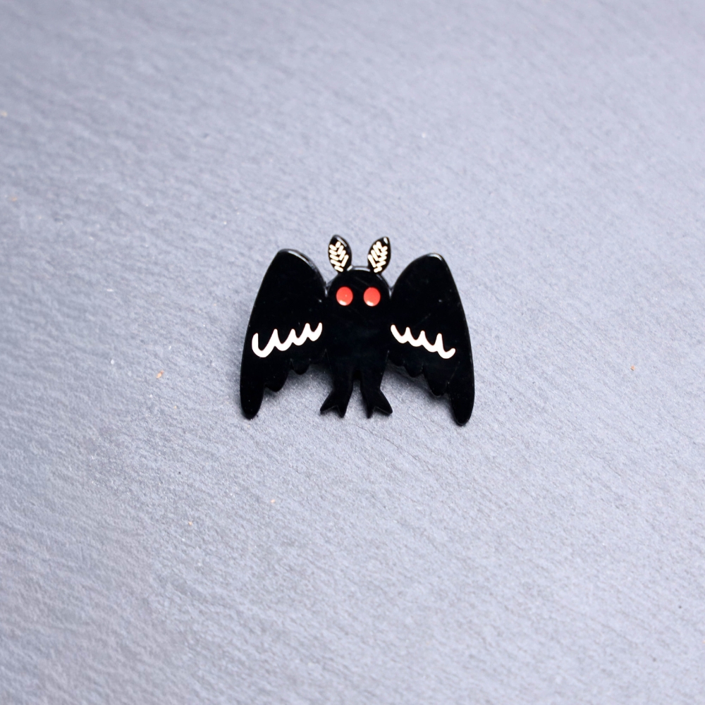 Mothman Big Wings Enamel Pin with black wings wide open and red eyes. These enamel pins are made with metal. These enamel pins are colorful, with a black backing.