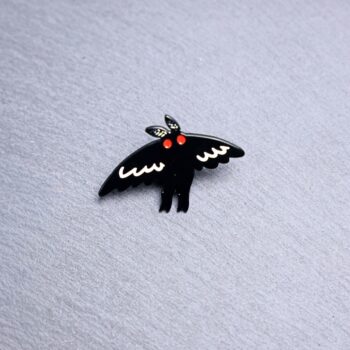 Mothman Flight Enamel Pin with wings in flight, black and white, and with red eyes. These enamel pins are made with metal. These enamel pins are colorful, with a black backing.