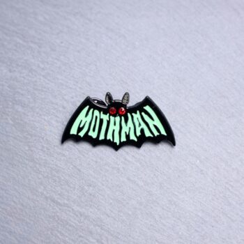 Mothman Green Enamel Pin says mothman on the front in green. Features red gel eyes. These enamel pins are made with metal. These enamel pins are colorful, with a black backing.