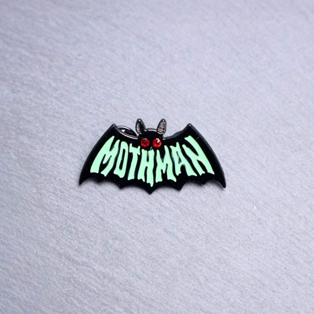 Mothman Green Enamel Pin says mothman on the front in green. Features red gel eyes. These enamel pins are made with metal. These enamel pins are colorful, with a black backing.
