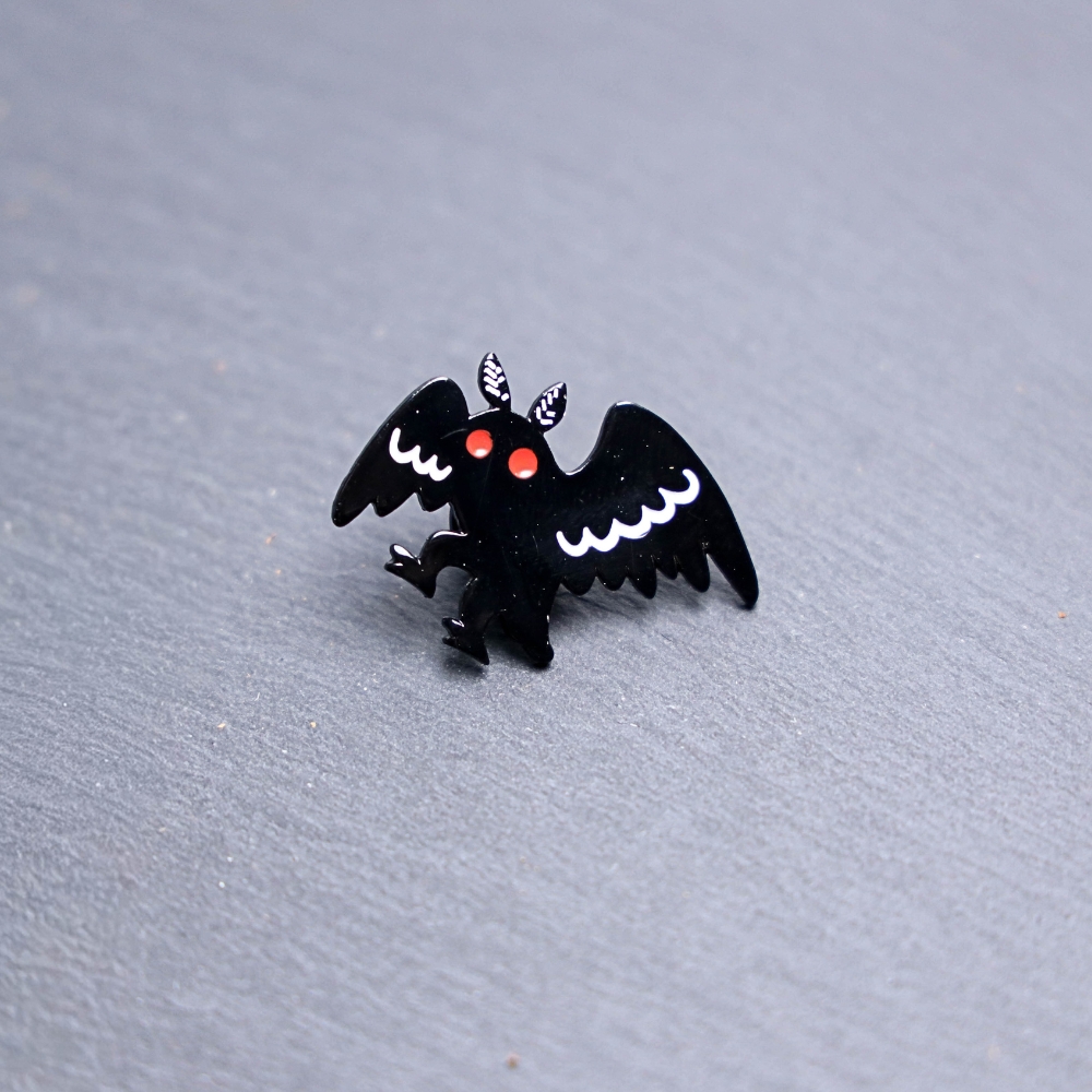 Mothman Walking Enamel Pin with black and white wings and red eyes. These enamel pins are made with metal. These enamel pins are colorful, with a black backing.