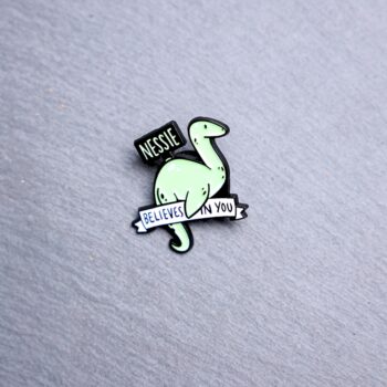 Nessie Believes In You Enamel Pin in green with sign. These enamel pins are made with metal. These enamel pins are colorful, with a black backing.
