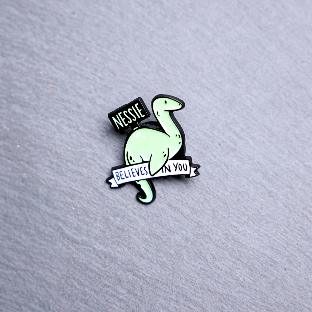 Nessie Believes In You Enamel Pin in green with sign. These enamel pins are made with metal. These enamel pins are colorful, with a black backing.
