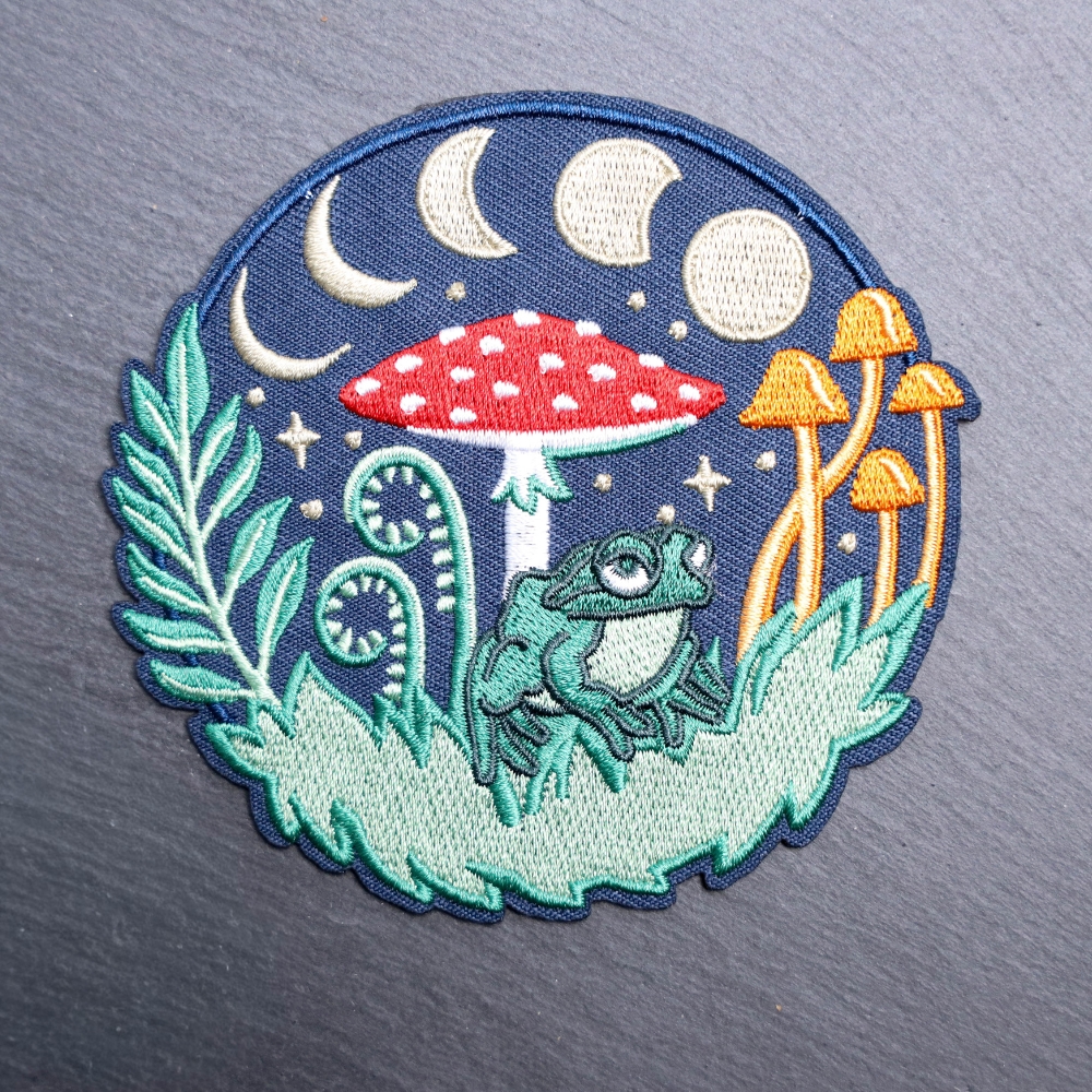 Forest Frog patch with a frog, mushrooms, and moons. Beautifully embroidered design with an adhesive for easy application to your favorite bag or article of clothing.