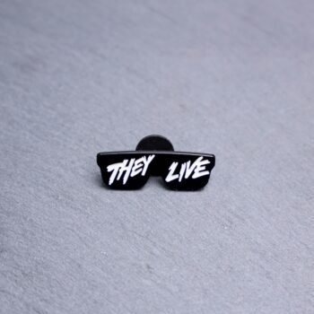 They Live Enamel Pin with black sunglasses and writing. These enamel pins are made with metal. These enamel pins are colorful, with a black backing.